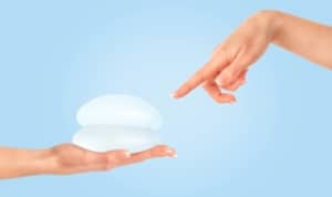 Female hand pointing at breast implants against a blue background, illustrating breast augmentation proportion.