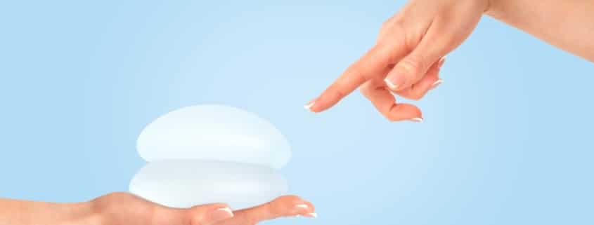 Female hand pointing at breast implants against a blue background, illustrating breast augmentation proportion.