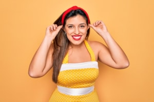 Playful woman in retro 1950s style smiling and pulling her ears with fingers against yellow background, concept of humor and hearing or audition issues