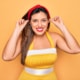 Playful woman in retro 1950s style smiling and pulling her ears with fingers against yellow background, concept of humor and hearing or audition issues