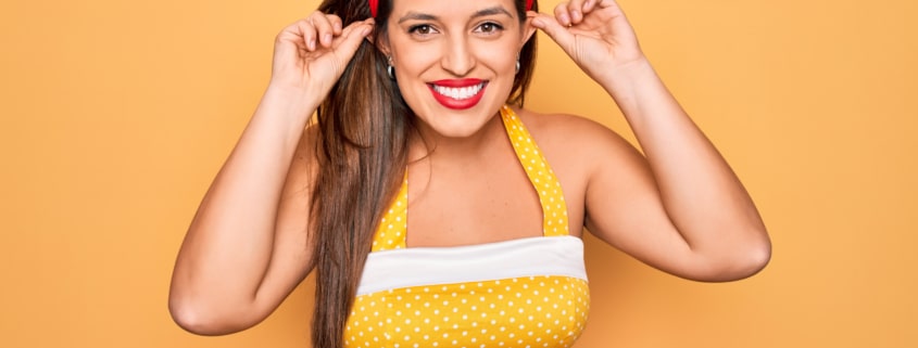 Playful woman in retro 1950s style smiling and pulling her ears with fingers against yellow background, concept of humor and hearing or audition issues