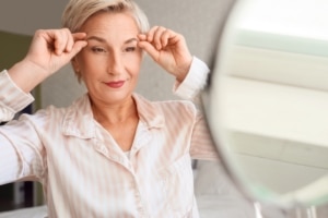 Mature woman performing face-building exercise near mirror in bedroom, concept of facial fitness and anti-aging skincare routine
