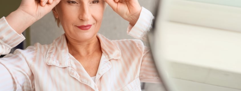 Mature woman performing face-building exercise near mirror in bedroom, concept of facial fitness and anti-aging skincare routine